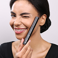 Eine Frau lächelt strahlend, während sie eine Mascara-Bürste in der Hand hält, und zeigt damit ihre Begeisterung für Schönheit und das Auftragen von Make-up.